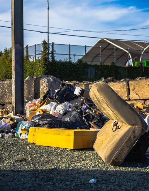 A pile of mixed waste materials is accumulated on a gravel surface beside a stone retaining wall. The collection includes black plastic rubbish bags, some partially torn open, revealing refuse inside, along with a large, battered beige car tire leaning against the rubbish. A yellow plastic container or toolbox rests on the ground among the debris. In the background, a metal pole supports overhead wiring, and behind the stone wall, there are green shrubs, a partially visible sports net or fencing, and a large white curved structure that appears to be a sports facility or shelter. The scene is outdoors under a bright blue sky with light clouds, suggesting daytime. The image presents an example of unmanaged or excess waste, illustrating the types of items that might require private rubbish removal services, as offered by Rubbish Removal Finchley, especially when alternative disposal options are unavailable or unsuitable in a private or on-site context.