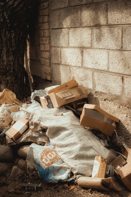 A pile of discarded cardboard boxes and paper debris, some flattened and others partially open, resting against a rough, concrete block wall outdoors. The cardboard varies in shades of brown, with printed labels and barcode stickers visible on some boxes. There are also crumpled brown paper and plastic packaging materials mixed in, creating a cluttered appearance. The pile is positioned on a surface of soil and small stones, with a large tree trunk on the left side of the image. The background features a weathered brick and concrete wall, suggesting an exterior environment where rubbish has been collected for removal. The scene conveys a typical example of waste accumulation suitable for private rubbish collection services such as those offered by Rubbish Removal Finchley, emphasizing the importance of proper disposal or clearance of unwanted packaging and packaging-related waste.