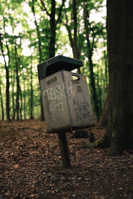 A weathered grey metal mailbox mounted on a black tilt-down post, located on a forest floor covered with brown soil and scattered leaves, with trees and dense green foliage in the background. The mailbox, which appears to be slightly tilted, has visible graffiti markings and faded stickers on its surface, and the lid is partially open. Surrounding the mailbox are tall tree trunks with rough, dark brown bark, and the dappled sunlight filters through the canopy above, creating a soft, natural glow. The scene suggests an outdoor setting where someone might leave correspondence or small packages, though the mailbox's condition and graffiti imply infrequent maintenance. The background indicates a peaceful woodland environment, contrasting with the practicality of private collection or waste disposal depending on how such objects are integrated into the landscape, which can relate loosely to concepts of alternative waste handling in remote or rural areas. Rubbish Removal Finchley’s expertise covers similar outdoor clearance tasks, often involving the proper disposal of items like this mailbox when it is no longer in use or needs removal as part of garden or property clearance services.