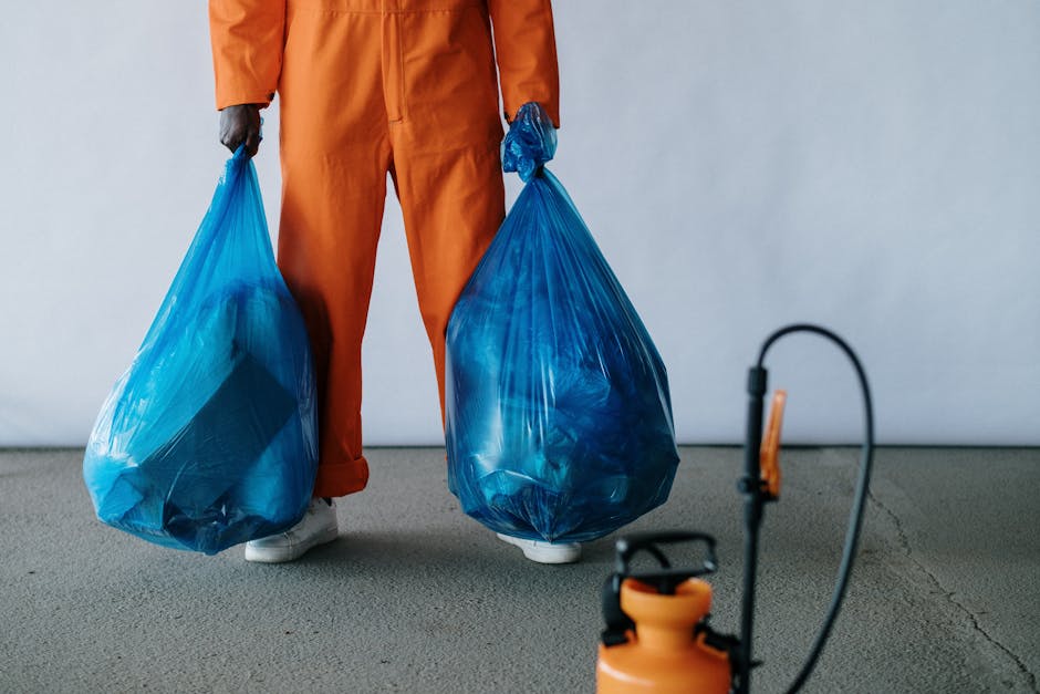 The image shows a person standing on a plain, light gray concrete floor, dressed in orange work overalls with matching gloves, holding two large blue plastic rubbish bags filled with waste materials. The bags are semi-transparent, revealing assorted waste inside, and are tightly knotted at the tops. The person's feet, wearing white shoes, are visible at the bottom of the image, with the bags hanging down on either side, close to their legs. In the background, a neutral white wall provides a simple backdrop. In the foreground, slightly out of focus, a small orange and black sprayer or hand-held cleaning device with a connected hose is situated on the floor, near the person's feet. This scene captures an on-site waste collection activity, reflecting a practical aspect of independent rubbish disposal, often associated with private waste removal services like those offered by Rubbish Removal Finchley, especially during circumstances where alternative waste management solutions are employed.