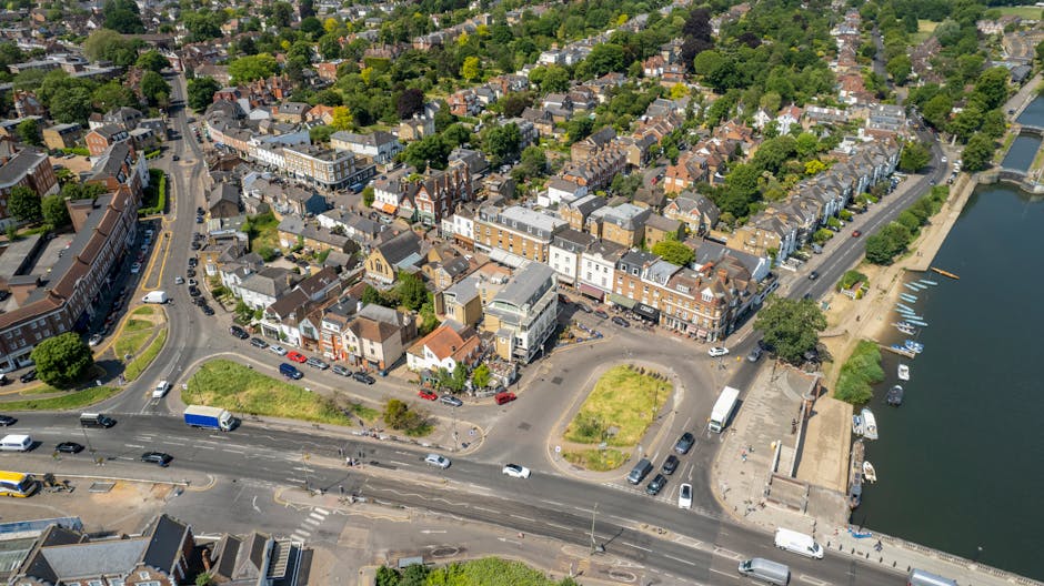 An aerial view of a small urban area showing a mix of commercial and residential buildings, with narrow streets and a prominent road running horizontally in the foreground. In the bottom right, there is a water body with several small boats docked along the quay. Adjacent to the water, a paved promenade features a stone wall and some trees providing shaded areas. The centre of the image includes a parking lot with various vehicles, such as cars, vans, and trucks, surrounding a small, landscaped green space with grass patches. The surrounding residential neighborhood extends into the background, characterized by closely packed houses with tiled or slate roofs, intersected by tree-lined streets and green areas. The overall scene is well-lit with daylight, capturing the typical environment where rubbish removal services might support property cleanouts or waste collection for both commercial and domestic clients, reflecting the context of private waste management in a busy town setting.