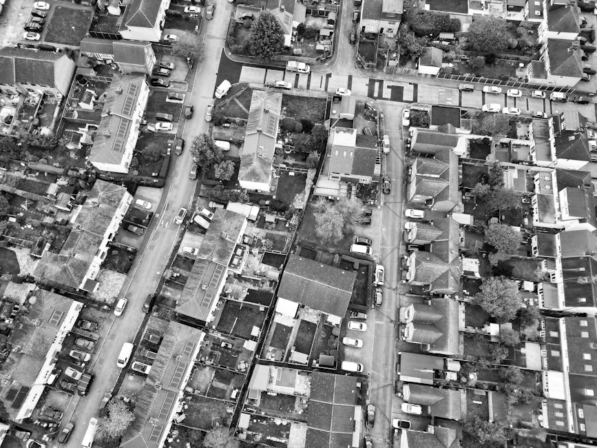 An aerial view of a residential urban area showing tightly packed houses with various roofing materials, including tiled and flat roofs, alongside narrow streets lined with parked cars in multiple spaces. Some properties have small gardens or yards, and a few trees are visible among the buildings. The streets are relatively congested with vehicles, and the overall scene is captured in black and white, highlighting the textures and shapes of rooftops, roads, and parked vehicles. This image illustrates a typical inner-city neighbourhood where private waste collection and rubbish removal services, like those provided by Rubbish Removal Finchley, are often used for house clearances or managing waste outside of municipal collection programs. The scene includes driveways and small outdoor areas consistent with houses in North Finchley, with the layout supporting on-site clearance and independent waste disposal solutions, relevant to the service category of rubbish removal.