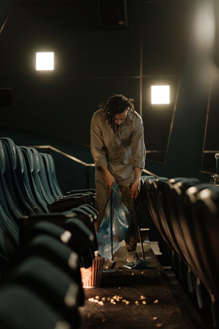 A person with dark hair tied back in a bun, dressed in a light grey jumpsuit, is seen in a dimly lit auditorium or cinema, leaning forward and collecting scattered trash from the dark carpeted floor. The individual is holding a large transparent plastic bag, partially filled, and is in the process of cleaning up debris, including small pieces of food and paper that are spread across the aisle. The surrounding environment features black, cushioned theatre seats aligned in rows along both sides of the aisle, with some seats visible in the foreground and background, and black walls with four rectangular lights emitting soft, white illumination. The scene suggests an on-site cleanup or rubbish removal operation, possibly conducted by Rubbish Removal Finchley in relation to waste clearance at a public venue like a cinema. The overall atmosphere is quiet and focused, with environmental lighting highlighting the person's activity in an indoor entertainment setting, emphasizing the importance of maintaining cleanliness through private rubbish collection services.