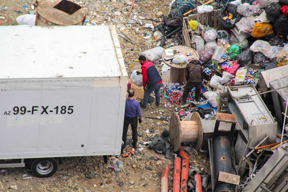 An outdoor scene depicts three individuals amid a large pile of mixed waste and debris on uneven ground. The waste includes black and white garbage bags, crumpled plastic packaging, cardboard, and discarded household items such as a wooden spool, metal pipes, and broken appliances. One person is wearing a red and black jacket, bending over as they handle waste, while another, dressed in dark clothing, is also engaged in sorting or lifting rubbish. A third individual with a purple shirt stands nearby, observing or assisting. A white vehicle with identification markings, including the code 'AZ 99-FX-185,' is parked at the edge of the scene, partially visible behind the waste. The environment appears to be a waste disposal or illegal dump site, with no visible structures or greenery, only dirt and scattered debris. The scene exemplifies the type of waste collection or clearance services offered by Rubbish Removal Finchley, highlighting the challenge of handling large volumes of varied rubbish typical of independent or private waste disposal efforts.