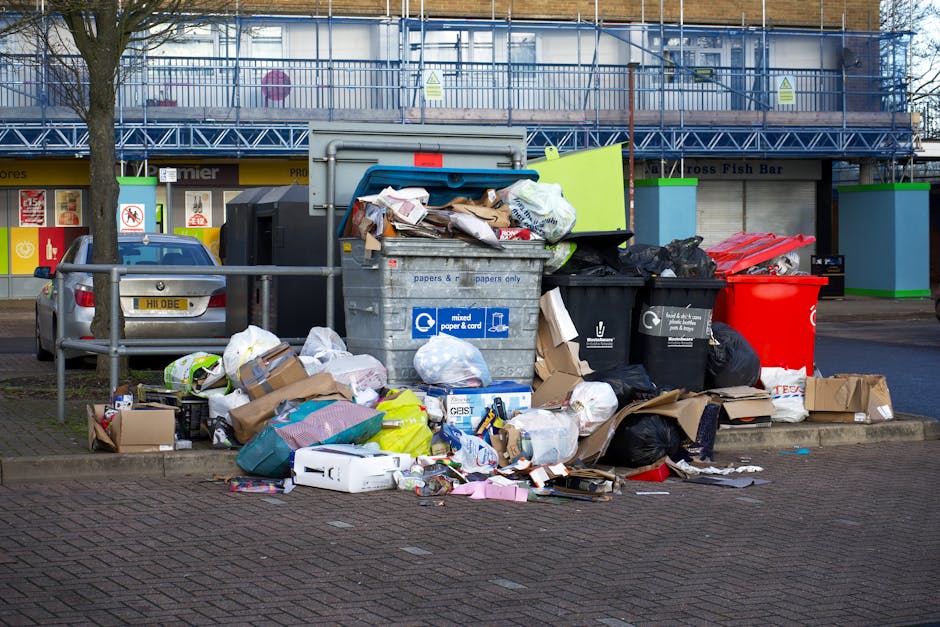 A large collection of mixed household waste, including cardboard boxes, plastic bags, paper, and discarded packaging, piled around and on top of multiple trash bins on a paved urban street. The waste appears to be overflowing, with some items spilling onto the surrounding pavement. The background shows a building with a blue scaffolding structure, indicating ongoing maintenance or construction work, and a row of commercial shops below, including a fish bar. A silver car is parked behind a metal railing on the left side of the scene. The area is illuminated by natural daylight, highlighting the disorganized state of the rubbish collection. This scene illustrates a scenario where independent rubbish disposal, such as private waste collection or on-site clearance services like Rubbish Removal Finchley, might be necessary due to access issues or overflowing bins.