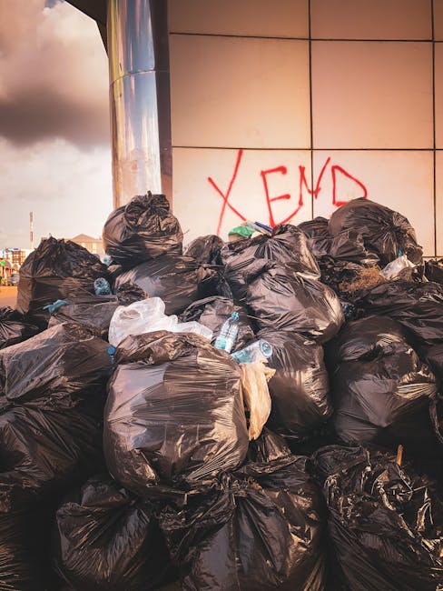 A large pile of black plastic rubbish bags is stacked against the side of a modern building with reflective tiles, some with visible creases and slight tears. The bags contain mixed waste, with a few transparent plastic bottles and pieces of discarded packaging partially protruding from the tops. The bags are haphazardly arranged, with some leaning and others resting on the pavement, indicating an ongoing or recent collection effort. Behind the pile, the reflective surface of the building catches the ambient light, creating a smooth, shiny finish in contrast to the textured trash. To the left, part of a street scene is visible with a cloudy sky overhead, suggesting an urban environment where private waste management services like Rubbish Removal Finchley might be employed for on-site clearance or alternative disposal arrangements. The graffiti with the word 'XEND' is spray-painted in red on the building's tiles, adding an element of urban context. Overall, this scene highlights the need for professional rubbish removal solutions for managing excess waste in city centers or restricted access locations, especially where traditional council collection may be limited.
